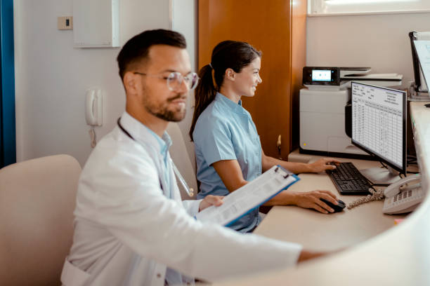 Doctors working at a hospital reception desk, reviewing patient data on a computer and clipboard.