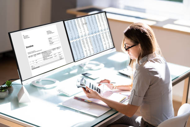 Person reviewing financial data and invoices on dual monitors with a calculator.