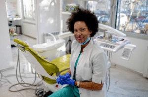 Smiling dentist in scrubs and gloves beside a dental chair in a bright office.