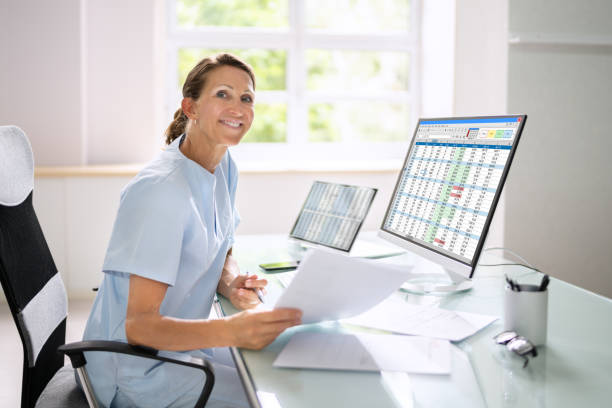 Smiling dental professional reviewing papers at a desk with financial data on screen.