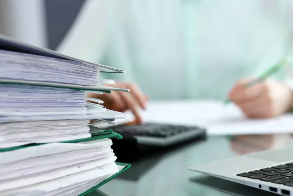 Stack of documents next to a person using a calculator and writing on paper.