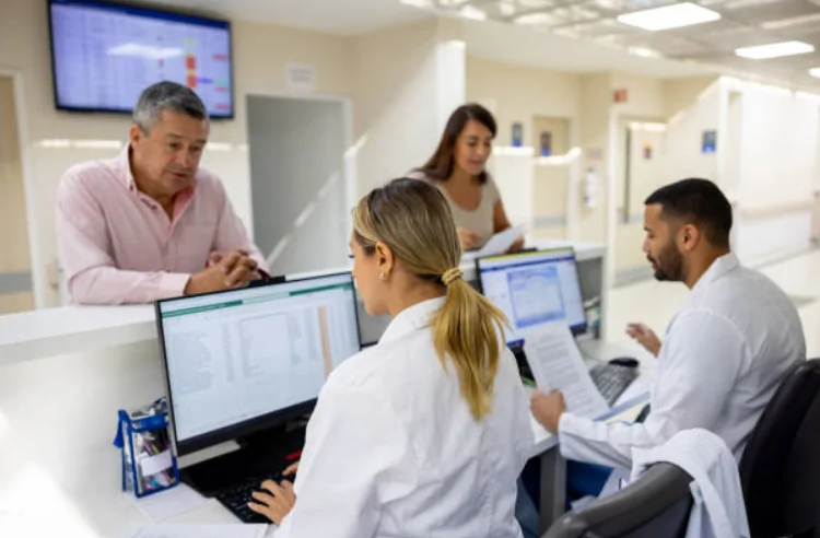 Medical staff working at a hospital reception desk assisting patients and reviewing records on computers.